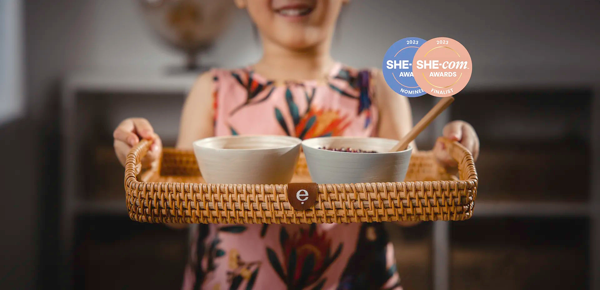 Girl holding a Montessori tray with two bowls, blurred background with 'SHE.com' logo.